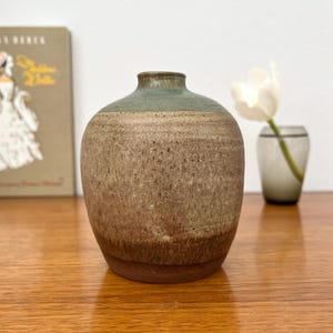 Può includere: Un vaso in ceramica fatto a mano, con un corpo arrotondato e un collo corto. Il vaso presenta una sfumatura di colori, che passa da una base marrone scuro a un centro marrone chiaro e una parte superiore verde-azzurro. Un fiore bianco in un piccolo vaso di vetro è sullo sfondo.