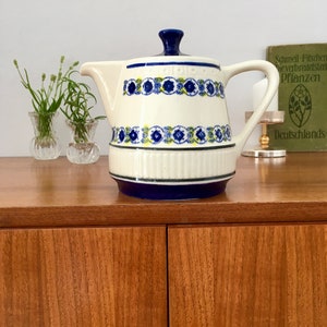 Op de afbeelding: Crèmekleurige keramische theepot met een blauw bloemmotief en een blauw deksel. De theepot heeft een handvat en een tuit. Het ontwerp bevat blauwe en gele bloemaccenten. De theepot staat op een houten oppervlak.