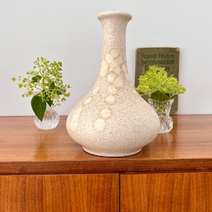Könnte beinhalten: Eine gesprenkelte beige Keramikvase mit rundem Boden und schmalem Hals. Die Vase hat eine strukturierte Oberfläche mit helleren kreisförmigen Mustern. Sie steht auf einem Holzschrank mit kleinen Glasvasen und Grünzeug.