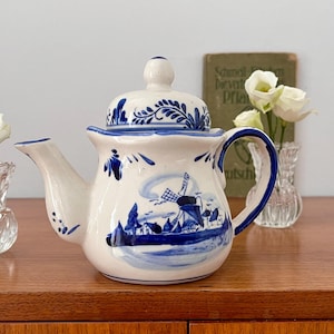 May include: A blue and white ceramic teapot with a windmill design. The teapot has a lid and a spout. It is sitting on a wooden table with a book and flowers in the background.