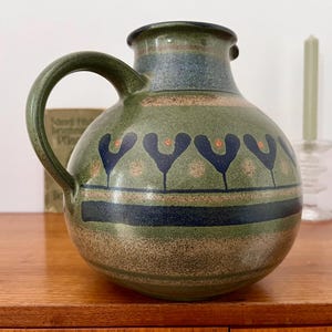 Könnte beinhalten: Eine grüne Keramikkanne mit blauem und braunem Design. Die Kanne hat eine breite Öffnung und einen Henkel. Sie zeigt ein sich wiederholendes Muster aus stilisierten Bäumen mit orangefarbenen Akzenten.