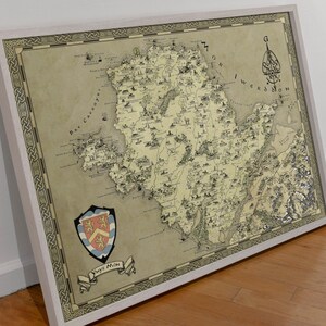 May include: A vintage-style map of a fictional island, with a coat of arms in the bottom left corner. The map is framed in a light wood frame. The map is titled "Ynys Mon" and features a compass rose in the top right corner.