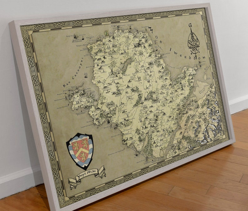 May include: A vintage-style map of a fictional island, with a coat of arms in the bottom left corner. The map is framed in a light wood frame. The map is titled "Ynys Mon" and features a compass rose in the top right corner.