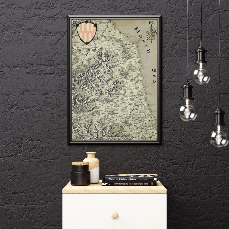 May include: A framed vintage map of a fictional land with a coat of arms in the top left corner. The map is in black and white with a beige background. The text "North Sea" is visible on the right side of the map. The map is hanging on a black wall with a small table underneath it.