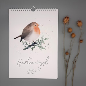 Könnte beinhalten: Ein Wandkalender mit einer Aquarellillustration eines Rotkehlchens auf einem grünen Zweig. Der Text "Gartenvögel 2027" ist in schwarzer Schreibschrift unter dem Vogel gedruckt. Der Kalender hat eine weiße Spiralbindung mit einer Aufhängung.