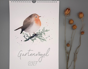 May include: A wall calendar featuring a watercolour illustration of a robin perched on a green branch. The text "Gartenvögel 2027" is printed in black script below the bird. The calendar has a white spiral binding at the top with a centre hanger.