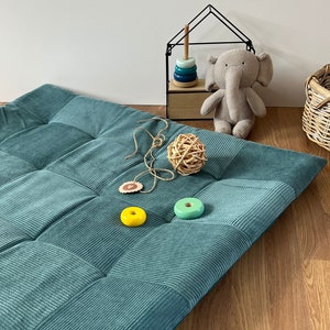 Può includere: Un tappetino da gioco trapuntato blu-verde con una superficie in velluto a coste testurizzata. Il tappetino è su un pavimento di legno con un piccolo scaffale di giocattoli in legno e un elefante di peluche sullo sfondo.