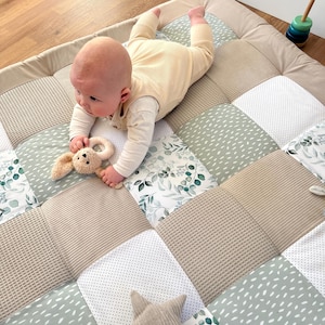 Puede incluir: Un bebé está acostado sobre una alfombra de juego de patchwork beige y verde. La alfombra presenta varias texturas y patrones, incluyendo diseños de gofres, lunares y florales. El bebé lleva un traje amarillo claro y sostiene un juguete de peluche de conejito.