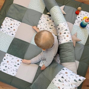 Peut inclure: Un tapis de jeu pour bébé en patchwork vert, beige et blanc. Le tapis présente différentes textures et motifs, dont du velours côtelé et des imprimés forestiers. Un boudin cylindrique et un oreiller sont également présents.