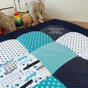 Może przedstawiać: Niebieska i biała patchworkowa mata do zabawy z wyszytym imieniem "Johann Levi" i datą "16.05.2022". Mata zawiera różne wzory, w tym gwiazdki, kropki, paski i nadruk z samochodami.