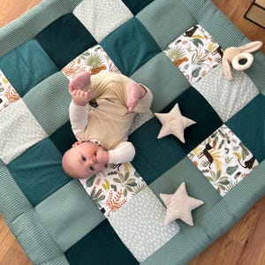 Pode incluir: Um bebê está deitado em um tapete de jogo de patchwork com quadrados de tecido waffle verde e tecido estampado com tema de selva. Duas almofadas em forma de estrela bege e um mordedor em forma de coelho estão por perto. O bebê usa um macacão bege.