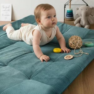 Könnte beinhalten: Ein Baby in cremefarbener Kleidung liegt auf einer tealfarbenen Cord-Spielmatte. Ein gelber Ring, ein Holzkugel und ein kleines Elefanten-Spielzeug befinden sich in der Nähe. Ein Schild im Hintergrund lautet "So fresh and wild and wonderful."