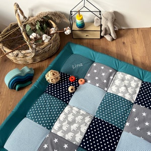 Op de afbeelding: Een patchwork speelmat met een teal rand en diverse patronen, waaronder sterren, olifanten en strepen. De naam "Lina" is op de mat geborduurd. Houten speelgoed en een rieten mand zijn in de buurt.