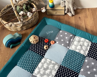 May include: A teal-bordered patchwork play mat featuring squares with star, elephant, and stripe patterns. The name "Lina" is embroidered on the mat. Wooden toys and a wicker basket are also visible.