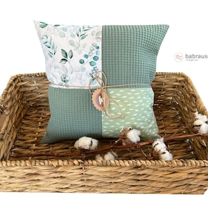 Può includere: Un cuscino con motivo verde e bianco con un fiocco di spago naturale si trova in un cesto di vimini intrecciato con boccioli di cotone. Il cuscino ha una texture a nido d'ape verde su un lato e uno sfondo bianco con foglie verdi e bacche blu sull'altro lato. Il cuscino è legato con un fiocco di spago naturale e un piccolo cartellino marrone con la scritta "babrause" su di esso.