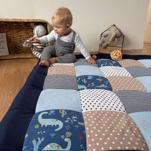 Op de afbeelding: Een patchwork speelmat met een marineblauwe rand en een verscheidenheid aan patronen in blauw, beige en wit. De patronen omvatten dinosaurussen, sterren, stippen en vichy. Een baby in grijze overalls zit op de mat.