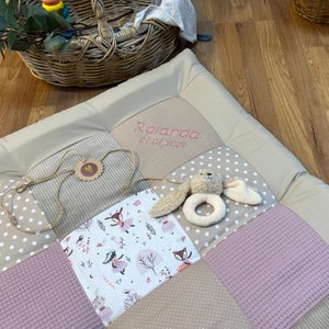 Könnte beinhalten: Ein beige-rosa Patchwork-Spielmatte für Babys mit dem Namen "Rolanda" und dem Datum "21.01.2024" in Rosa gestickt. Die Matte weist verschiedene Stoffmuster auf, darunter Punkte, Streifen und einen Waldtierdruck. Ein Plüsch-Hasenrassel ist befestigt.