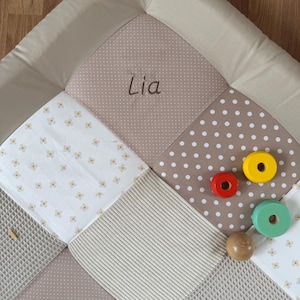 Könnte beinhalten: Eine quadratische Spielmatte mit Patchwork-Design aus beigen, weißen und braunen Stoffbahnen. Die Matte enthält Abschnitte mit weißen Blumenmustern, weißen Punkten auf braunem Grund und einer Waffelstruktur. Der Name Lia ist in Braun eingestickt.