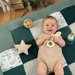 Könnte beinhalten: Ein lächelndes Baby in einem beigen Strampler liegt auf einer Patchwork-Spielmatte. Die Matte hat grüne, weiße und gemusterte Quadrate. Das Baby hält eine beige Hasenrassel und ist von einem sternförmigen Kissen, einem Holzkugel und einem Stapelspielzeug umgeben.