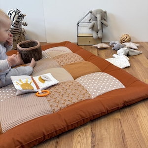 Może przedstawiać: Patchworkowa mata do zabawy dla dziecka z rdzawą obwódką i różnymi kwadratami materiału w neutralnych odcieniach. Na macie wyhaftowano imię "Leonard". Obecne są zabawki dla dzieci, w tym książeczka, gryzak i pluszaki.