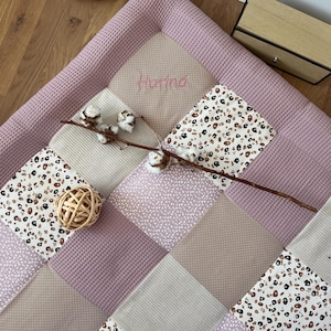 May include: A pink and beige patchwork play mat with a waffle texture border. The mat has a leopard print pattern, a small floral pattern, and a solid beige fabric. The name "Hanna" is embroidered in pink on the mat.