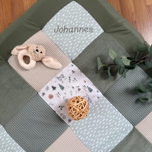 Puede incluir: Una alfombra de juego para bebés de patchwork en tonos verdes, beige y blanco. La alfombra presenta una variedad de texturas y patrones, incluyendo tejido de gofre y tela de lunares. Un sonajero de conejito de peluche y una bola tejida están sobre la alfombra. El nombre "Johannes" está impreso.