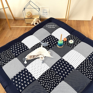 Może przedstawiać: Mata do zabawy w kolorze granatowym i białym z różnymi wzorami, w tym kotwicami, gwiazdami i kratą. Mata ma spersonalizowany napis "Leandro 17.05.2022". Mata jest również ozdobiona drewnianą zabawką do układania i pluszową zabawką.