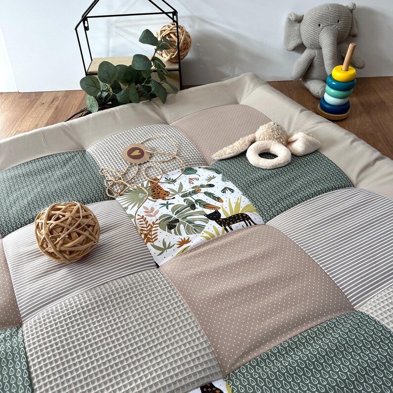 May include: A patchwork baby play mat with various textures and patterns, including waffle weave, stripes, and polka dots. The mat features a jungle-themed panel with animal illustrations and tropical foliage. Additional toys include a wooden ball, a stuffed elephant, and a ring stacker.