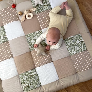 Może przedstawiać: Dziecko leży na beżowo-zielonej patchworkowej macie do zabawy z zabawką w kształcie gwiazdki i zabawką w kształcie króliczka. Mata wykonana jest z różnych tkanin o różnych wzorach, w tym grochy, paski i wzory kwiatowe.