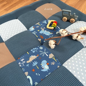 Op de afbeelding: Een patchwork babyspeelmat met een blauw wafelontwerp. De mat heeft vierkanten van blauwe, witte stippen en dinosaurussenstof. Een houten speelgoedtrekker, een houten ringspeelgoed en een takje katoen liggen op de mat. De naam "Luis" is geborduurd.