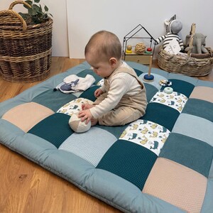 Op de afbeelding: Een gewatteerde speelmat met een patchworkontwerp van blauwgroene, lichtblauwe en beige vierkanten, voorzien van gestructureerde wafelstof en katoen met walvispatronen. De mat bevat een kleine ronde bal en diverse speeltjes.