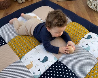 May include: A square patchwork play mat featuring a variety of fabric patterns including navy blue with white polka dots, mustard yellow with rainbows, white with whale illustrations, and beige with small dots, arranged in a grid layout.