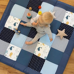 Könnte beinhalten: Eine Patchwork-Spielmatte für Babys in Blautönen mit Quadraten aus Punkten, Streifen und Walmotiven. Ein Baby sitzt auf der Matte mit Holzspielzeug und sternförmigen Kissen. Die Matte hat einen dunkelblauen Rand.
