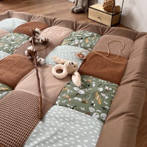 Può includere: Un tappeto gioco patchwork con una varietà di tessuti e fantasie, tra cui velluto a coste, tessuto a nido d'ape e stampe floreali nei toni del marrone, verde e beige. Un coniglietto di peluche e un ramo di cotone sono sul tappeto.