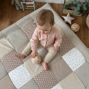 Könnte beinhalten: Eine quadratische Baby-Spielmatte mit einem Patchwork-Design aus beigen, cremefarbenen und weißen Stoffquadraten mit Waffelstrukturen, Punkten und kleinen Blumenmustern. Die Matte besteht aus mehreren gesteppten Abschnitten in neutralen Farbtönen.