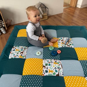 Könnte beinhalten: Eine Patchwork-Spielmatte mit einem tealfarbenen Rand und bunten Quadraten. Die Quadrate weisen Muster wie weiße Sterne auf Marineblau, gelbe Punkte und Safari-Tierdrucke auf. Ein Baby sitzt auf der Matte mit Holzspielzeug.