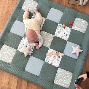 May include: A green, quilted baby play mat with a patchwork design. The mat features squares of solid green, blue stripes, and floral patterns. A baby is lying on the mat, with a star-shaped pillow and a plush bunny toy nearby. A small red ring and wooden toy are also on the mat.