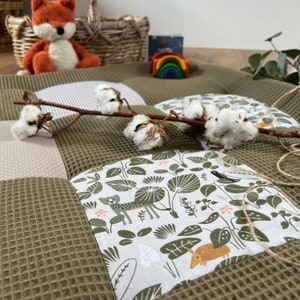 Może przedstawiać: Zielona i biała wzorzysta mata do zabawy o różnych fakturach. Mata przedstawia zwierzęta z dżungli, w tym geparda, ptaka i pancernika. Mata jest idealna do zabawy dla niemowląt i małych dzieci.
