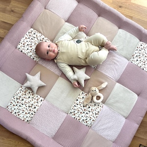 Könnte beinhalten: Eine rosa und beige Patchwork-Spielmatte mit einem darauf liegenden Baby. Die Matte hat verschiedene Texturen und Muster, darunter Punkte, Leopardenmuster und Streifen. Außerdem befinden sich zwei Plüschtiere auf der Matte, ein Stern und ein Hase.