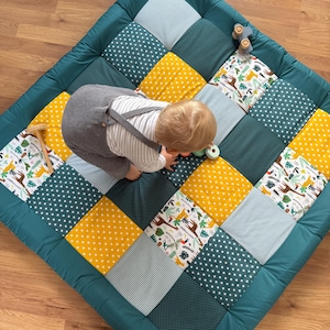 Op de afbeelding: Een gewatteerde speelmat met een patchwork ontwerp met vierkanten van teal, gele, witte en blauwe stoffen. Sommige vierkanten hebben witte sterren, stippen of een safari dierenprint. Een kleine houten speelgoedauto ligt op de mat.