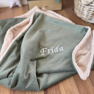 Könnte beinhalten: Eine salbeigrüne Corddecke mit einem weichen, beigefarbenen Sherpa-Futter. Die Decke ist mit dem Namen "Frida" in weißer Schrift bestickt. Die Decke ist gefaltet und zeigt die Textur des Cordes und des Sherpas.