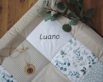 May include: A beige and white patchwork baby play mat with the name "Luano" in black script. The mat features various patterned squares, including polka dots, waffle texture, and a floral design. A sprig of eucalyptus and a small tag are included.