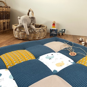 Puede incluir: Una alfombra de juego acolchada azul y beige con un patrón de ballena y arcoíris. La alfombra está sobre un suelo de madera con una cesta de mimbre, un elefante de peluche y un camión de juguete de madera.
