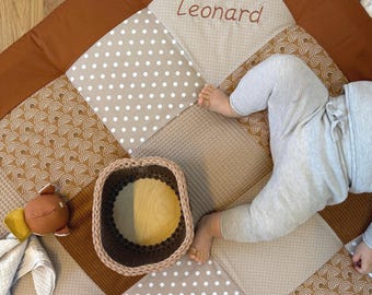 May include: A patchwork baby play mat with brown, tan, and white squares. The mat features a woven basket, a stuffed animal, and the name "Leonard" embroidered in brown. A baby is lying on the mat.