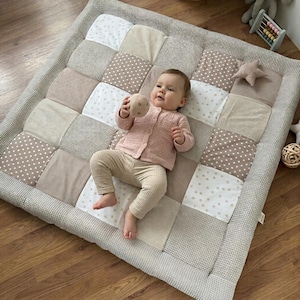 May include: A patchwork baby play mat in neutral tones of beige, cream, and taupe. The mat features a variety of fabric patterns, including polka dots and stripes. A baby in a pink cardigan and beige pants is lying on the mat, holding a toy.