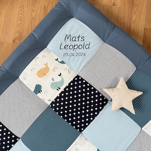 May include: A blue patchwork baby play mat with various patterns, including whales, rainbows, polka dots, and stripes. The mat is personalised with the name "Mats Leopold" and the date "29.04.2026". A star-shaped cushion in beige is placed on the mat.