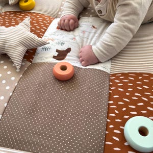 Könnte beinhalten: Ein Baby spielt auf einer mehrfarbigen Spielmatte mit einem braunen, weißen und orangen Farbschema. Die Matte hat verschiedene Texturen und Muster. Das Baby spielt mit Holzringen in Orange und Hellblau.