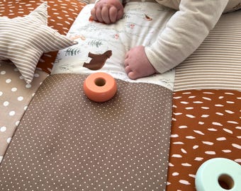 May include: A baby plays on a multi-colored play mat with a brown, white, and orange color scheme. The mat has different textures and patterns. The baby is playing with wooden rings in orange and light blue.
