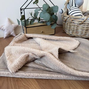 Könnte beinhalten: Eine beige Decke mit einem Cord-Außenmaterial und einem weichen, flauschigen Innenfutter. Die Decke ist gefaltet und zeigt die kontrastierenden Texturen des gerippten Stoffes und des Plüschfutters. Die Decke ist wahrscheinlich für ein Baby oder Kind.