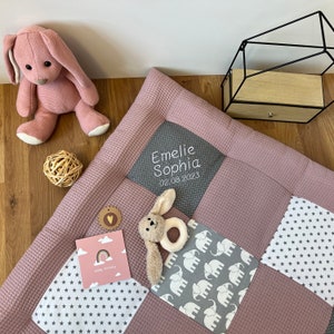 Könnte beinhalten: Eine rosa-graue Baby-Spielmatte mit einem weißen gestickten Namen "Emelie Sophia" und dem Geburtsdatum "02.08.2023". Die Matte hat verschiedene Muster, darunter Sterne, Elefanten und ein graues Quadrat mit weißen Punkten. Ein kleines Stofftier-Häschen mit einem Beißring liegt auf der Matte.